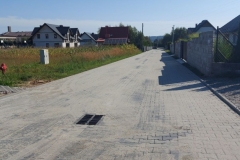 Droga z kostki brukowej. Po obu stronach drogi domy jednorodzinne. W oddali Szkoła podstawowa w Rzuchowej.