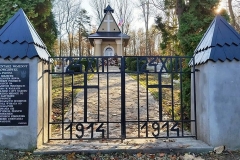 Metalowa brama pomiędzy betonowymi kolumnami, w tle kaplica cmentarna