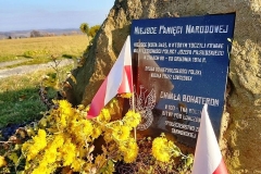 Pamiątkowa tablica przytwierdzona do skały, przed nią żółte kwiaty i biało-czerwona flaga