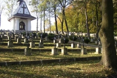Mogiły żołnierskie z metalowymi krzyżami na tle kaplicy cmentarnej