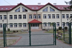 Frontowa ściana budynku szkoły z wejściem, na pierwszym planie zielona brama