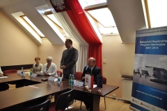 Wójt przemawia w pozycji stojącej na tle biało-czerwonej flagi, z prawej strony granatowy baner