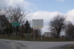 Pośrodku tablica informacyjna dotycząca inwestycji, z lewej strony zielony znak z nazwą miejscowości
