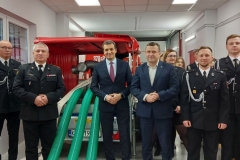 Wójt ze strażakami obok przyczepy, z której wystają trzy zielone węże strażackie