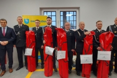Wójt ze strażakami, którzy trzymają nowy sprzęt schowany w czerwonych pokrowcach