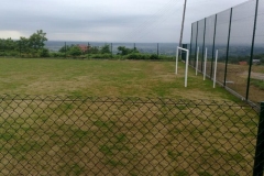 Kawałek siatki od ogrodzenia, za nią boisko, po prawej biała bramka do piłki nożnej