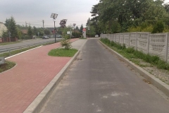 Z lewej chodnik z czerwonej kostki brukowej, pośrodku wąska droga asfaltowa, z prawej betonowy mur