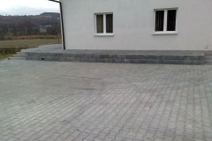 Plac z kostki brukowej obok bocznej ściany budynku, na ścianie dwa okna