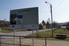 Tablica turystyczna w Janowicach, za nią droga asfaltowa i zabudowania