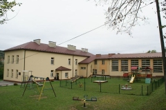 Plac zabaw dla dzieci, za nim budynek szkoły i sala gimnastyczna