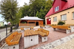Betonowo-drewniane ławeczki przed drewnianą wiatą pokrytą dachem z blachy, z prawej pomarańczowy budynek remizy strażackiej pokryty czerwonym dachem