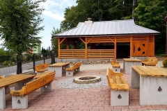 Betonowo-drewniane ławeczki przed drewnianą wiatą pokrytą dachem z blachy