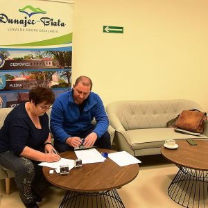 Moment podpisywania umowy przy okrągłym stoliku, na ścianie plakat LGD Dunajec-Biała