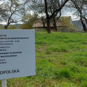 Tabliczka z informacją o dofinansowaniu. W tle widoczna droga asfaltowa i sad.