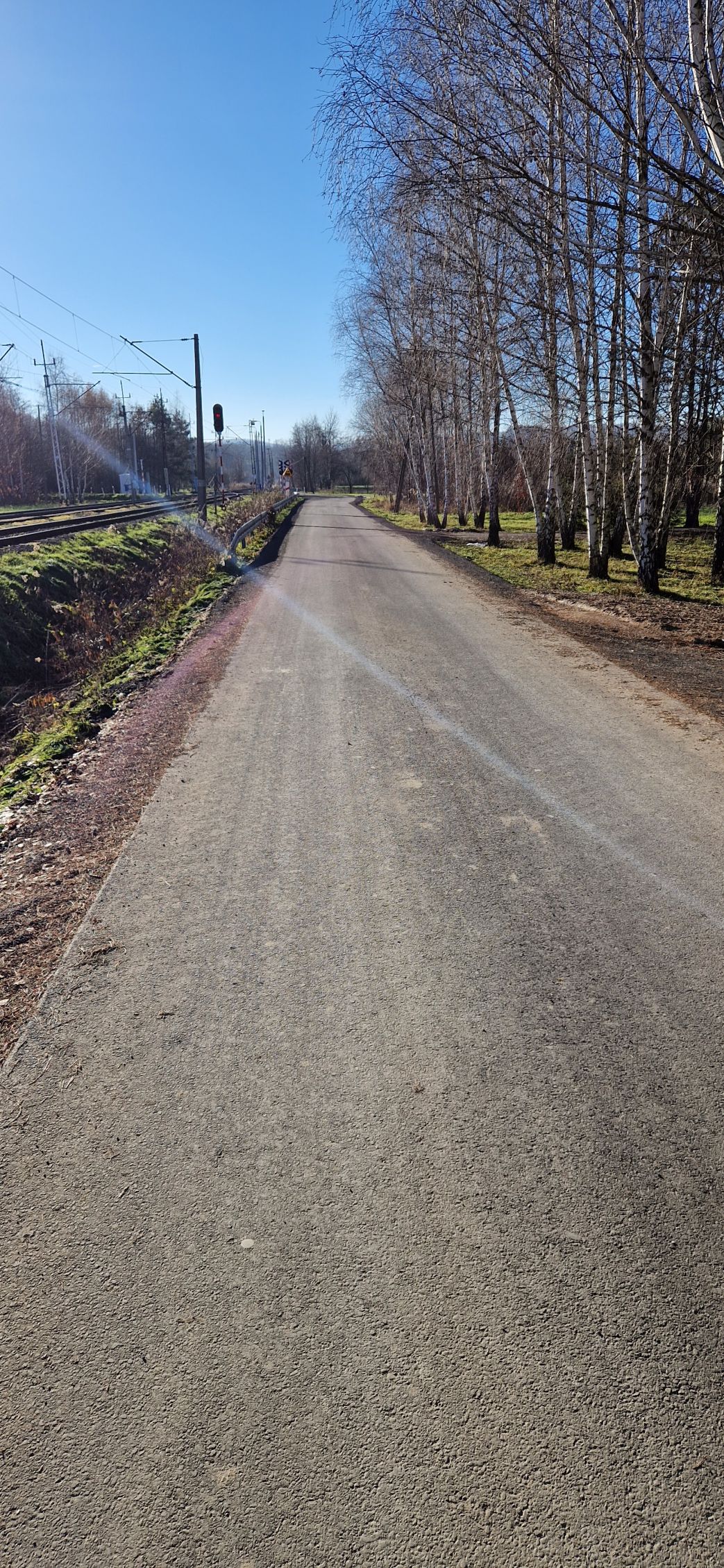 asfaltowy odcinek drogi wzdłuż torów, z prawej strony rosną drzewa