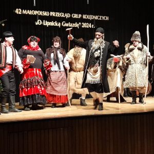 Grupa kolędników tańczy i śpiewa na scenie. Na pierwszym planie postacie żyda i pastucha.