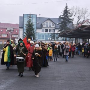 Grupa osób maszeruje przez plac. Na czole pochodu dzieci przygrywają na instrumentach.