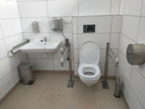 Toaleta przystosowana dla potrzeb osób z niepełnosprawnością. Po prawej stronie biała miska ustępowa. Po obu stronach pochwyty. Po lewej stronie umywalka. Nad umywalką pojemnik na mydło i suszarka.