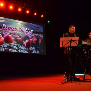 Na zdjęciu dwie osoby stoją na scenie przed mikrofonami i pulpitami na nuty. W głębi sceny na ścianie wyświetlony jest obraz przedstawiający tulipany. Nad obrazem świecą się światłem czerwonym reflektory.