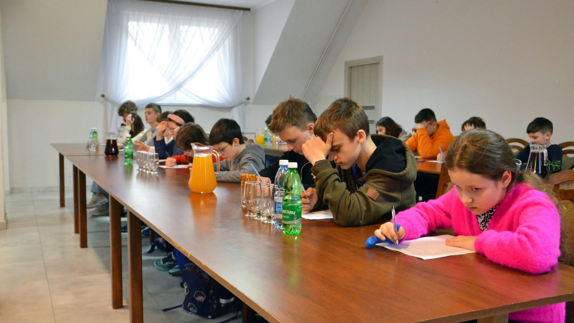 Na zdjęciu dzieci siedzą przy stołach, w rękach trzymają długopisy i rozwiązują test.