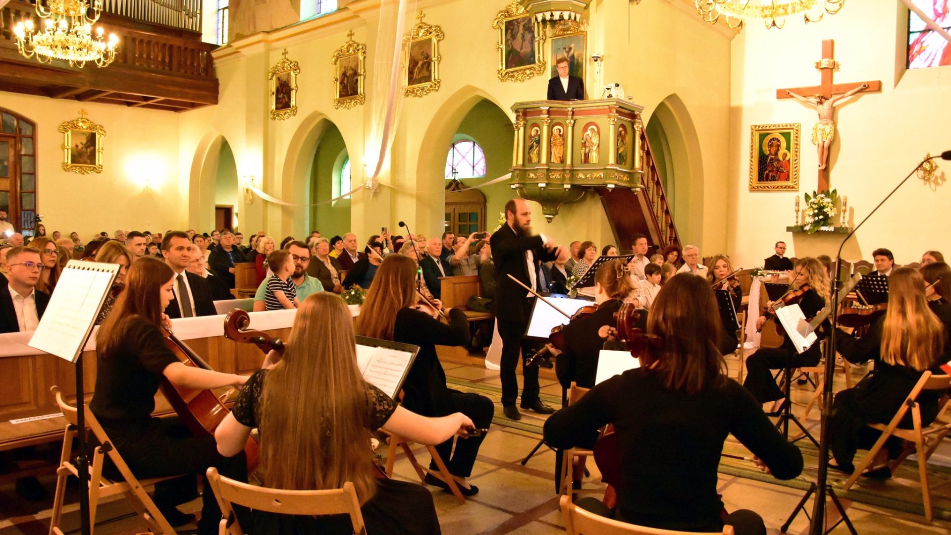 Na zdjęciu orkiestra wraz z dyrygentem daje koncert w kościele. Na drugim planie publiczność w ławach kościelnych.