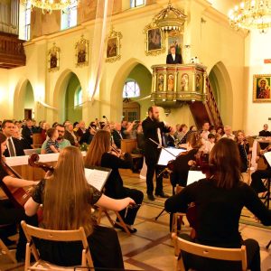 Na zdjęciu orkiestra wraz z dyrygentem daje koncert w kościele. Na drugim planie publiczność w ławach kościelnych.