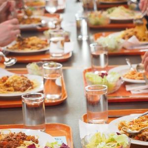 Tacki ze spaghetti w stołówce, przy każdej szklanka z wodą. Dłonie trzymające sztućce w trakcie posiłku.