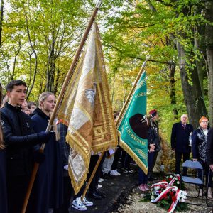 Młodzież stojąca w rzędzie trzyma sztandary. Po prawej kilka osób stoi na baczność.
