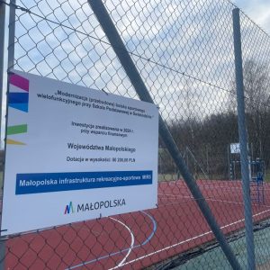 Boisko wielofunkcyjne. Na pierwszym planie ogrodzenie (siatka), na której jest zamontowana tablica informująca o przyznanym dofinansowaniu.