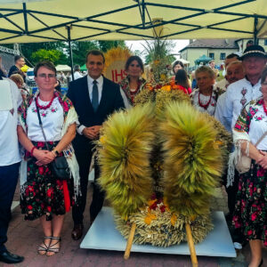 Grupa osób w strojach ludowych stoi wokół wieńca dożynkowego.