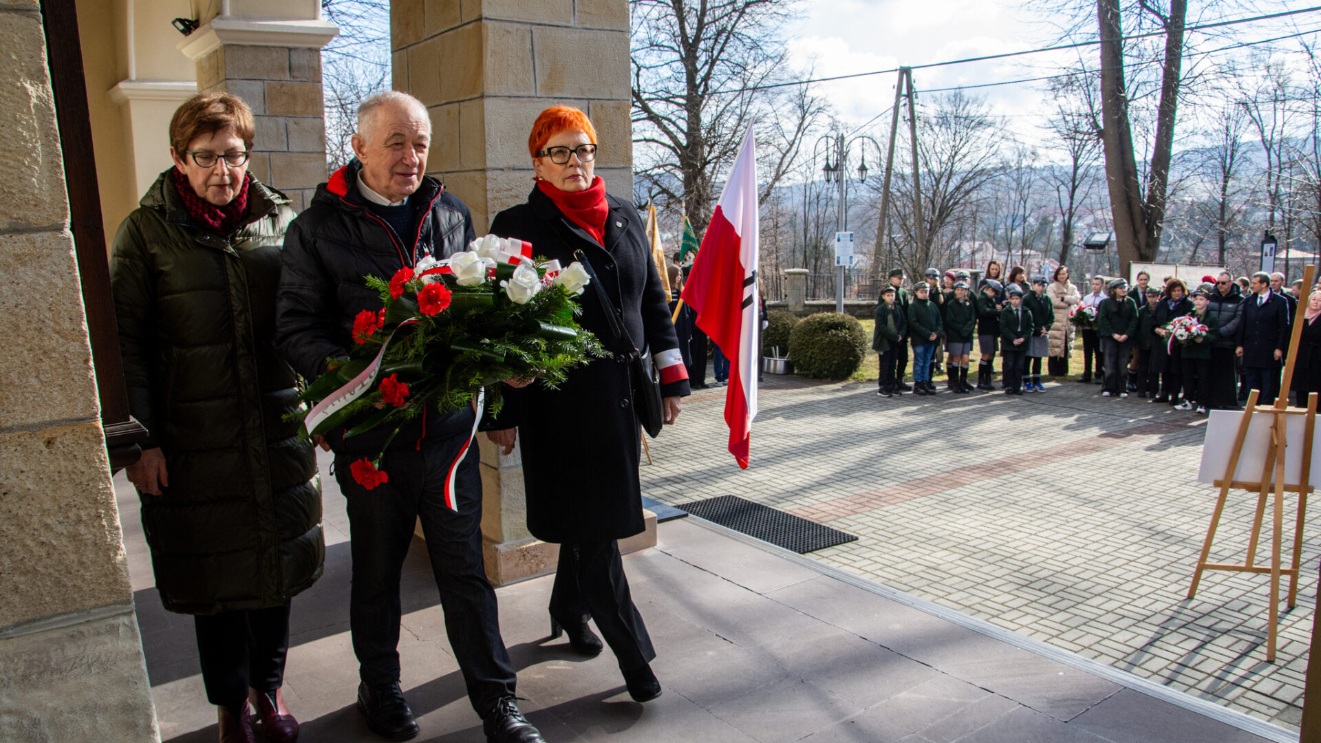 Na zdjęciu dwie kobiety i jeden mężczyzna, który niesie wiązankę kwiatów.