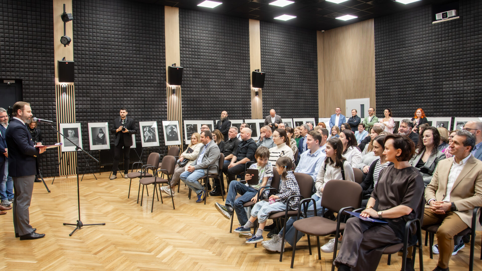 Grupa osób w dużej sali siedzi na krzesłach. Jeden mężczyzna przemawia do mikrofonu.