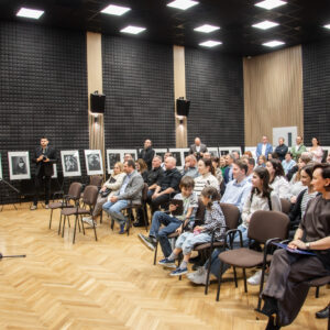 Grupa osób w dużej sali siedzi na krzesłach. Jeden mężczyzna przemawia do mikrofonu.