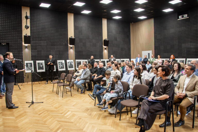 Grupa osób w dużej sali siedzi na krzesłach. Jeden mężczyzna przemawia do mikrofonu.