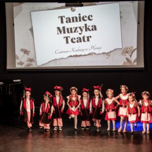 Dzieci w strojach krakowskich stoją na scenie, po lewej opiekunka/ W tle duży ekran z napisem Taniec Muzyka Teatr